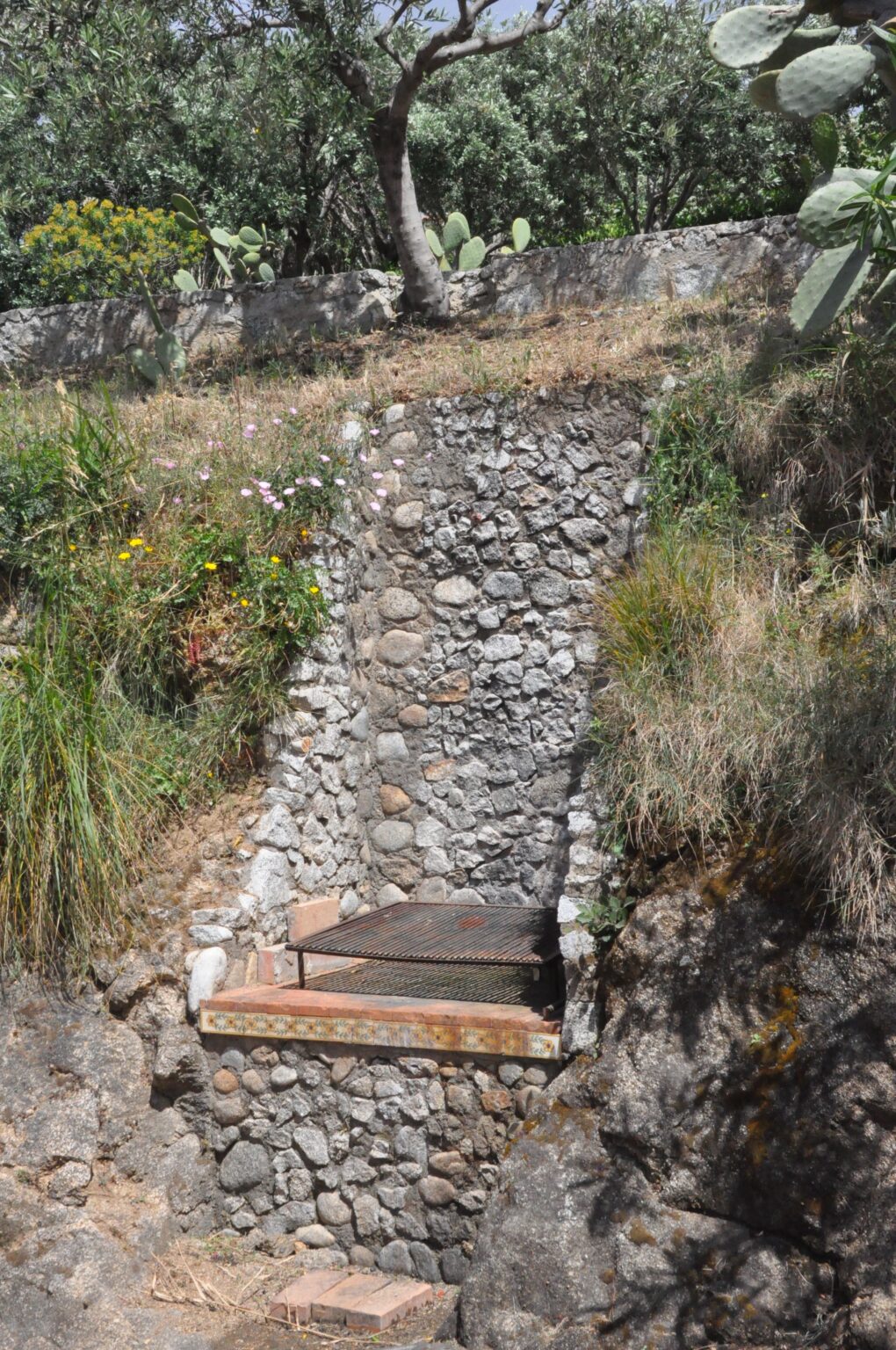 Stone wall with a metal grill set into a hillside, surrounded by greenery and wildflowers, showcasing a rustic outdoor cooking area. | Only on SopranoVillas