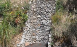 Stone wall with a metal grill set into a hillside, surrounded by greenery and wildflowers, showcasing a rustic outdoor cooking area.