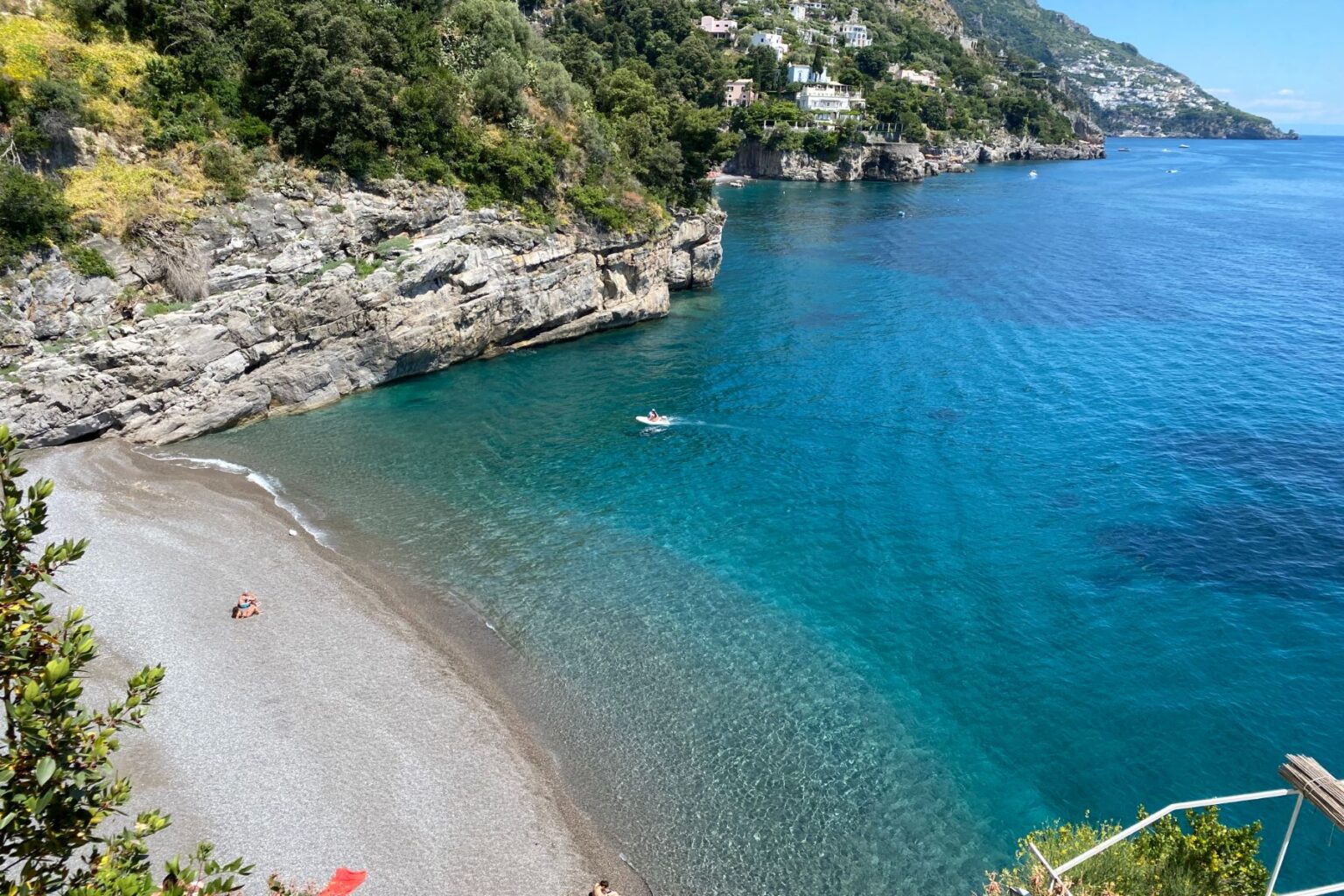 Scenic coastal view featuring a serene beach with clear turquoise waters, rocky cliffs, and lush greenery along the shoreline. A small boat is seen on the water, highlighting the tranquil atmosphere of the location. | Only on SopranoVillas