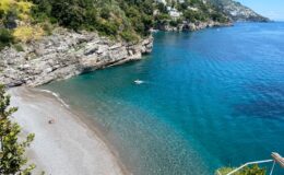Scenic coastal view featuring a serene beach with clear turquoise waters, rocky cliffs, and lush greenery along the shoreline. A small boat is seen on the water, highlighting the tranquil atmosphere of the location.