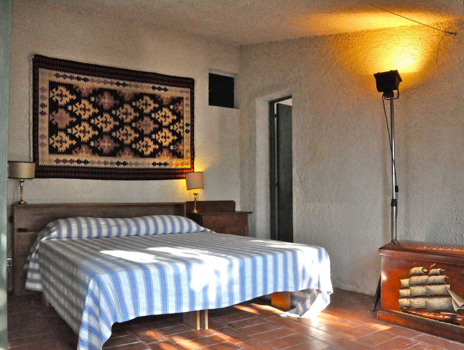 Cozy bedroom featuring a striped blue and white bedspread, wooden furniture, a decorative wall tapestry, and ambient lighting from a floor lamp. The room has a warm and inviting atmosphere, ideal for relaxation. | Only on SopranoVillas