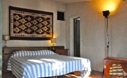 Cozy bedroom featuring a striped blue and white bedspread, wooden furniture, a decorative wall tapestry, and ambient lighting from a floor lamp. The room has a warm and inviting atmosphere, ideal for relaxation.