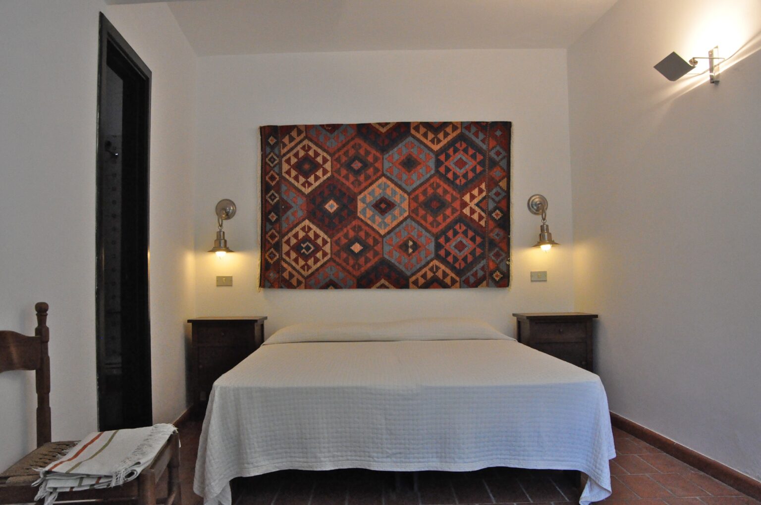 Cozy bedroom featuring a white bedspread, decorative wall tapestry with geometric patterns, and warm lighting from wall sconces. The room includes wooden nightstands and a rustic chair, creating a welcoming and stylish atmosphere. | Only on SopranoVillas