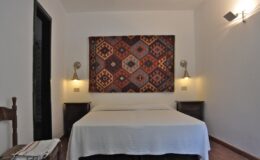 Cozy bedroom featuring a white bedspread, decorative wall tapestry with geometric patterns, and warm lighting from wall sconces. The room includes wooden nightstands and a rustic chair, creating a welcoming and stylish atmosphere.