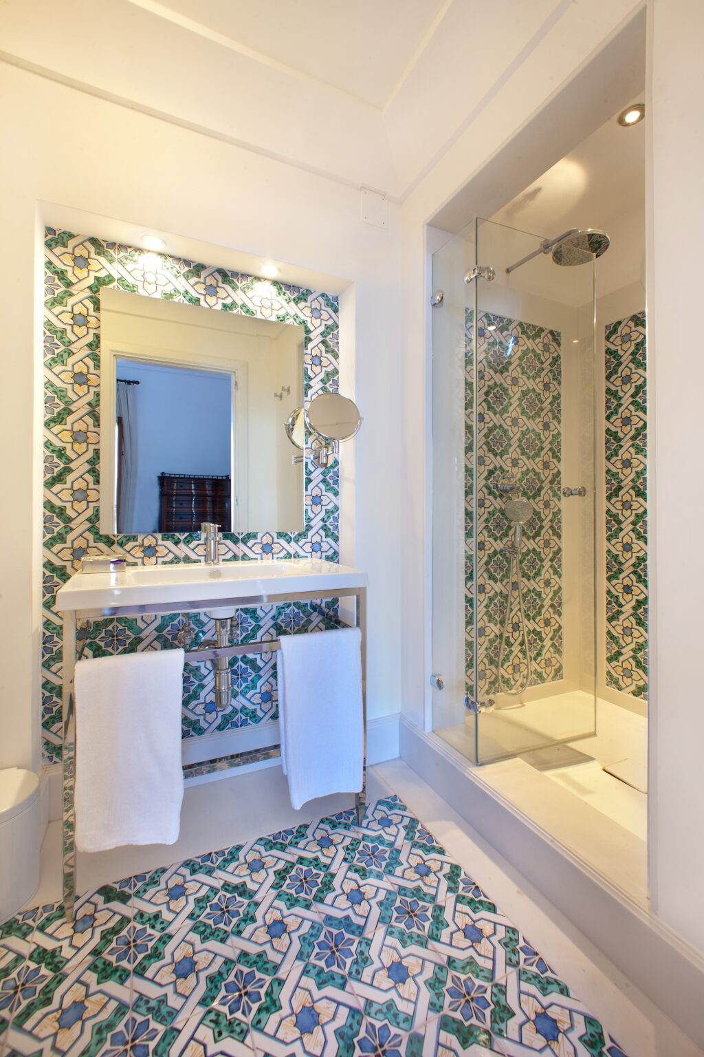 Modern bathroom featuring a stylish vanity with a large mirror, decorative tiled walls in blue and green patterns, and a glass shower enclosure. Soft white towels hang neatly, enhancing the clean and inviting atmosphere. | Only on SopranoVillas
