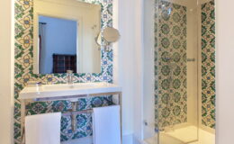 Modern bathroom featuring a stylish vanity with a large mirror, decorative tiled walls in blue and green patterns, and a glass shower enclosure. Soft white towels hang neatly, enhancing the clean and inviting atmosphere.