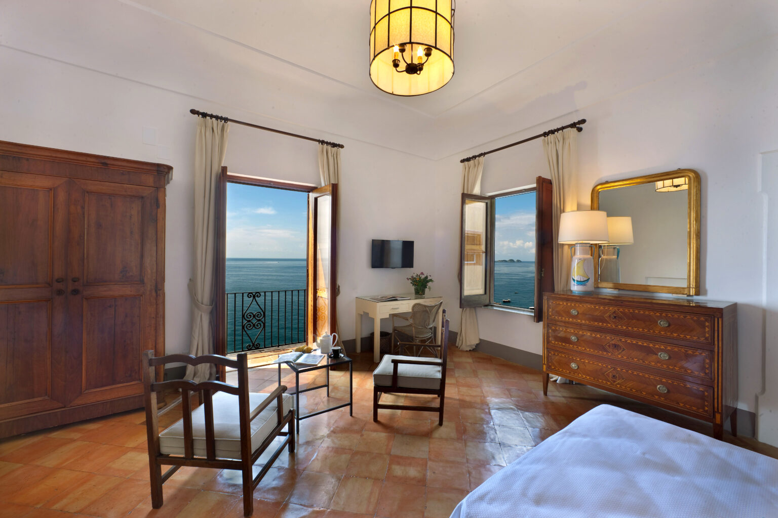 Cozy hotel room featuring large windows with ocean views, wooden furniture, a stylish chandelier, and a comfortable seating area, ideal for relaxation and enjoying the scenery. | Only on SopranoVillas