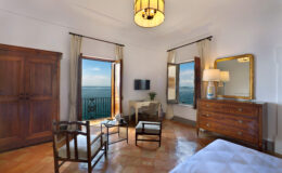 Cozy hotel room featuring large windows with ocean views, wooden furniture, a stylish chandelier, and a comfortable seating area, ideal for relaxation and enjoying the scenery.