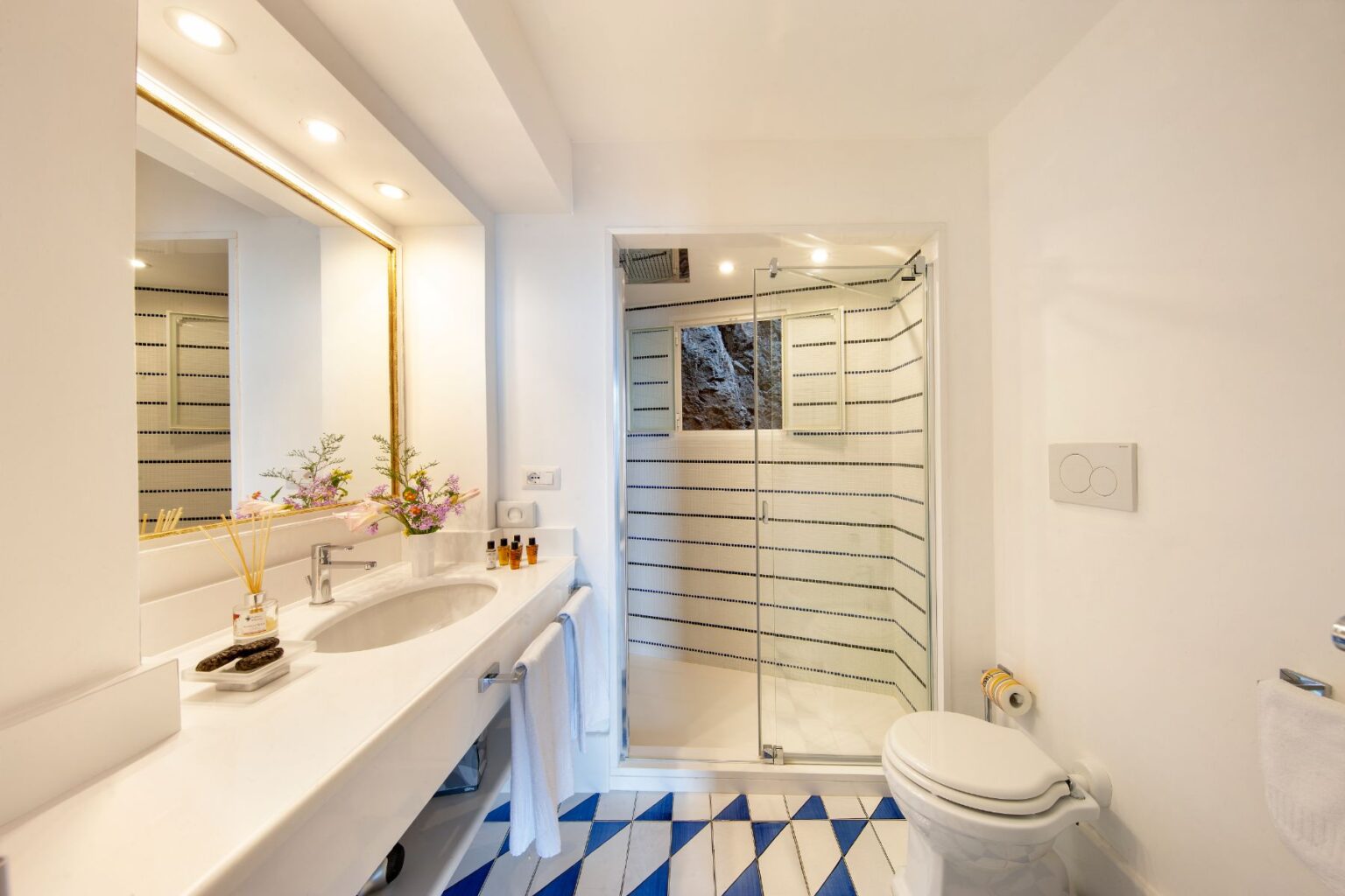 Modern bathroom featuring a spacious vanity with a large mirror, stylish sink, and organized toiletries. The glass shower enclosure showcases a contemporary design, while the blue and white patterned tiles add a vibrant touch to the floor. Natural light brightens the space, enhancing the clean and elegant aesthetic. | Only on SopranoVillas