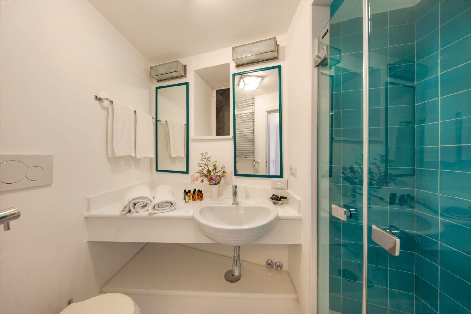 Modern bathroom featuring a sleek design with a glass shower, minimalist sink, and stylish mirrors. The turquoise tiles add a vibrant touch, while the decor includes subtle floral accents for a fresh look. | Only on SopranoVillas