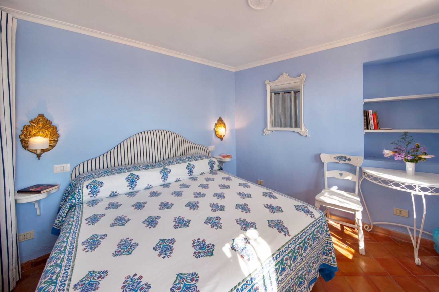 Cozy blue-themed bedroom featuring a patterned bedspread, elegant wall sconces, a small writing desk, and a shelf with books, perfect for relaxation and comfort. | Only on SopranoVillas
