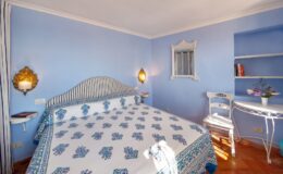 Cozy blue-themed bedroom featuring a patterned bedspread, elegant wall sconces, a small writing desk, and a shelf with books, perfect for relaxation and comfort.