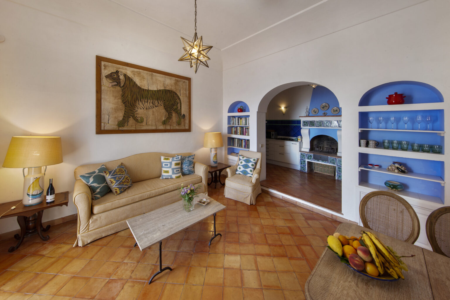 Cozy living room featuring a beige sofa with colorful cushions, a wooden coffee table, and a decorative tiger painting on the wall. The space includes a lamp and a bookshelf, with an archway leading to a kitchen area adorned with blue tiles and decorative ceramics. The room has terracotta flooring and a dining table with a bowl of fresh fruit. | Only on SopranoVillas
