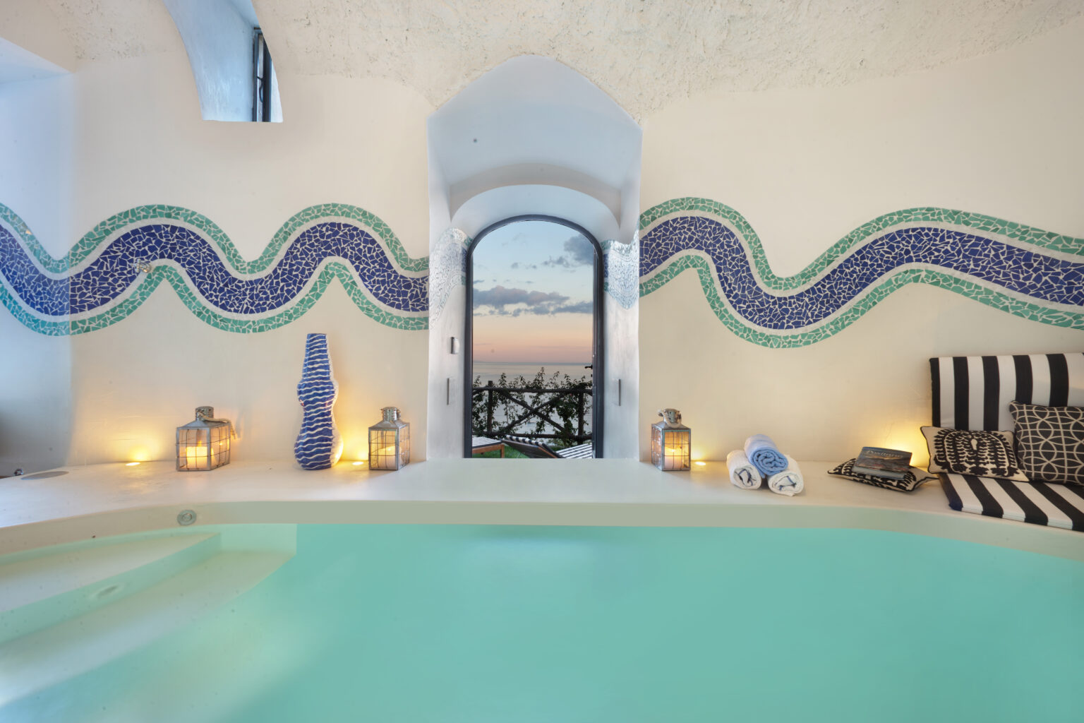 Luxury villa interior in Italy featuring a serene swimming pool, mosaic wall designs, and stunning views through an arched window, perfect for an upscale getaway. | Only on SopranoVillas