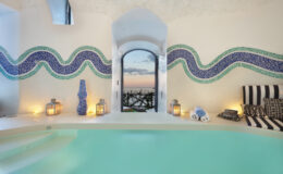 Luxury villa interior in Italy featuring a serene swimming pool, mosaic wall designs, and stunning views through an arched window, perfect for an upscale getaway.