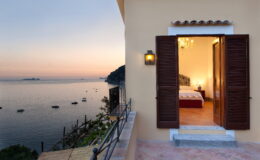 Luxury villa for rent in Italy featuring a stunning sunset view from the terrace, with open shutters revealing a beautifully decorated bedroom. Ideal for a serene getaway by the coast.
