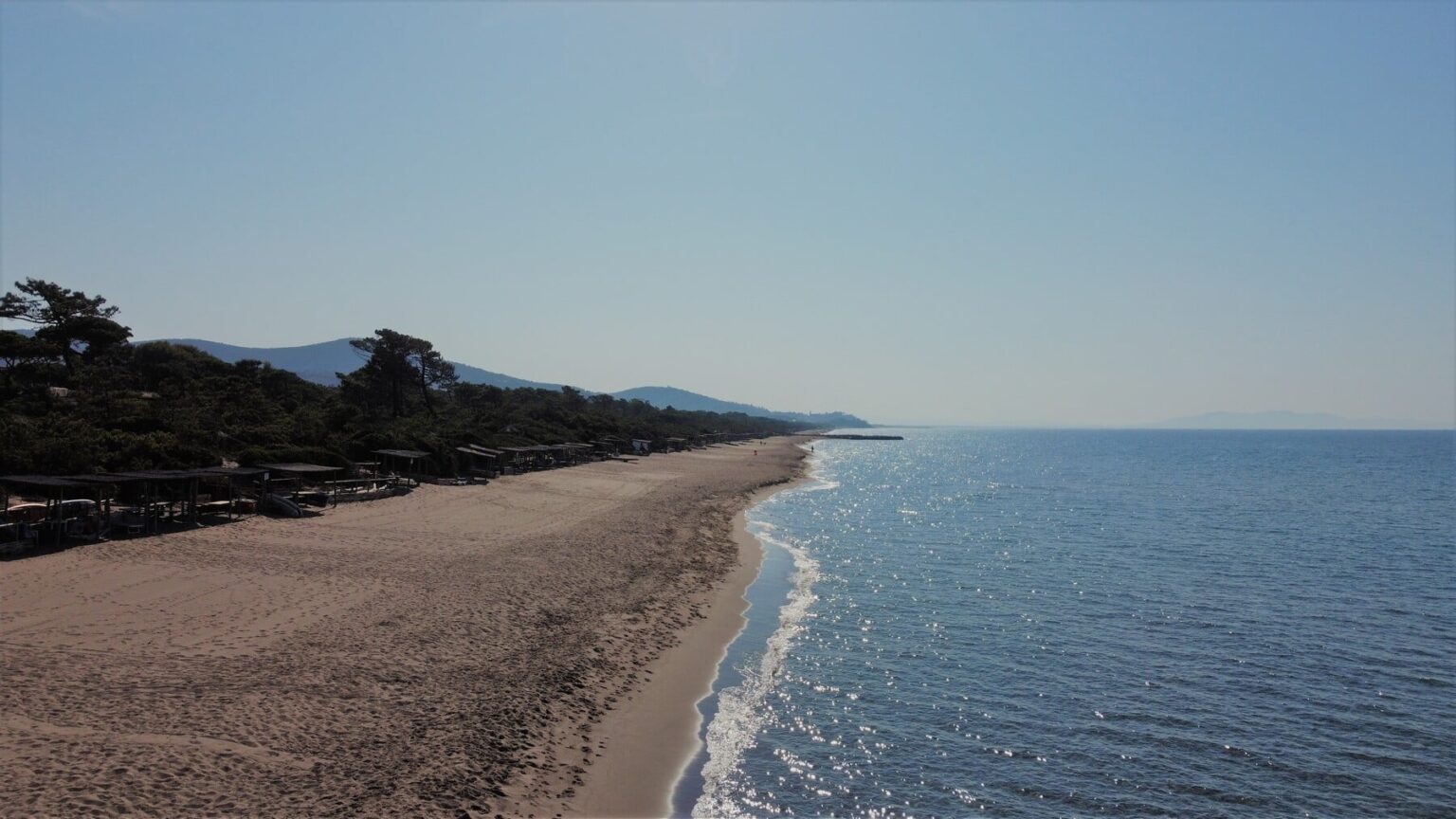 A serene beach with golden sand, gentle waves lapping at the shore, and distant mountains under a clear blue sky. Sunlight sparkles on the water, creating a tranquil atmosphere ideal for relaxation and leisure activities. | Only on SopranoVillas