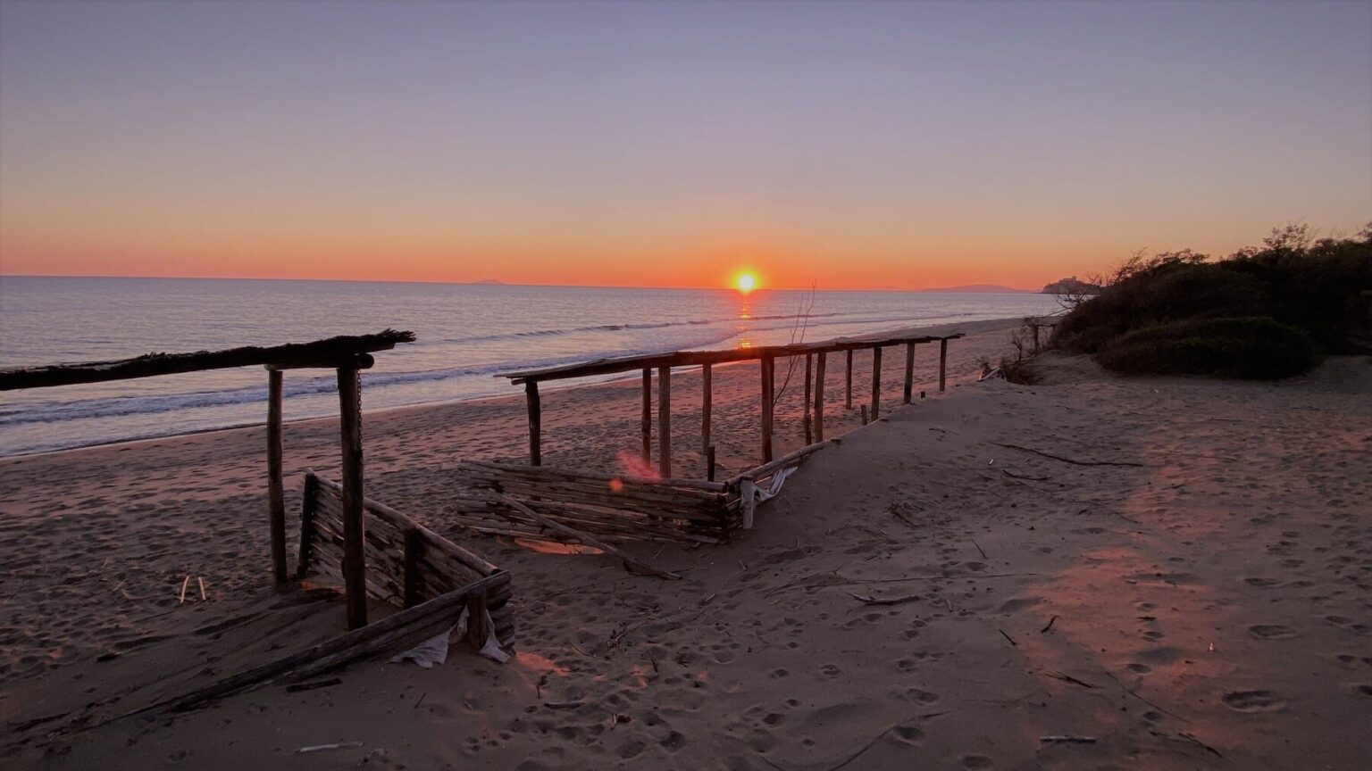 Sunset over a tranquil beach with wooden structures on the sand, creating a serene coastal atmosphere. Gentle waves lap at the shore as the sky transitions to warm hues of orange and purple. | Only on SopranoVillas