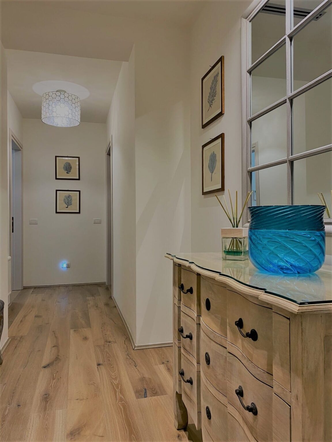 Bright hallway interior featuring framed leaf artwork, wooden flooring, and a decorative blue glass vase on a console table. The space exudes a modern and inviting atmosphere. | Only on SopranoVillas