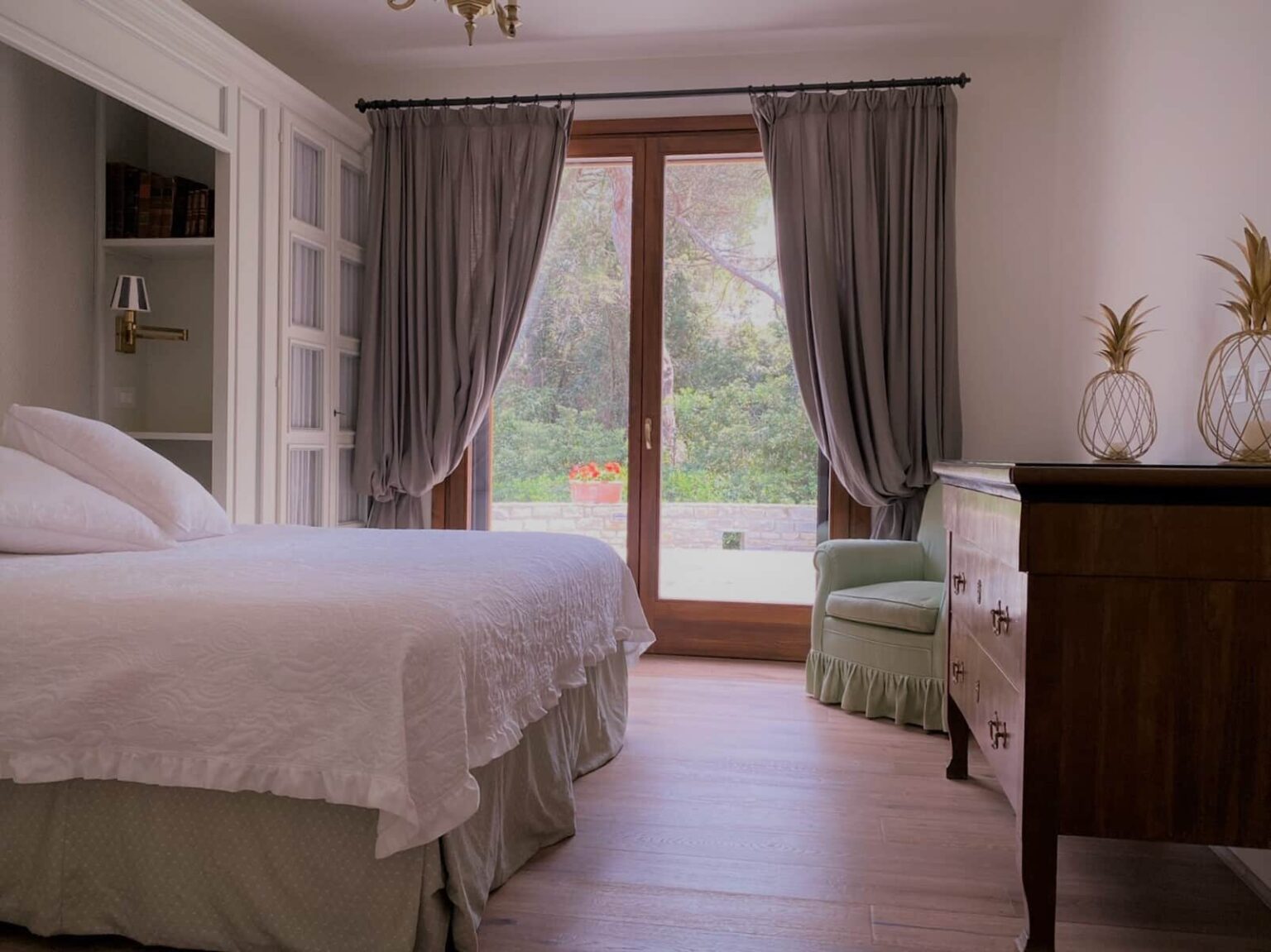 Cozy bedroom interior featuring a neatly made bed with white bedding, a wooden dresser, and a green accent chair. Large windows with gray curtains provide natural light and a view of the garden outside. Ideal for relaxation and comfort in a serene setting. | Only on SopranoVillas