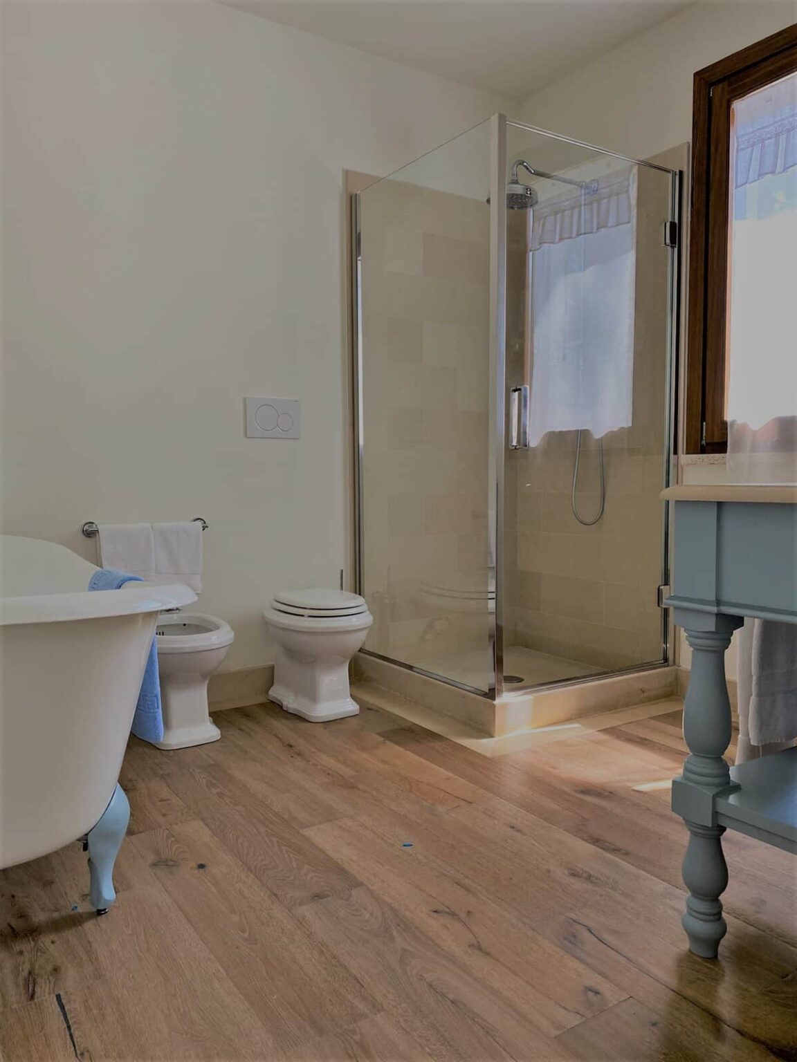 Modern bathroom featuring a glass shower enclosure, a freestanding bathtub, and white fixtures. The space is designed with natural wood flooring and includes a light blue cabinet, towels, and a window with sheer curtains for natural light. | Only on SopranoVillas