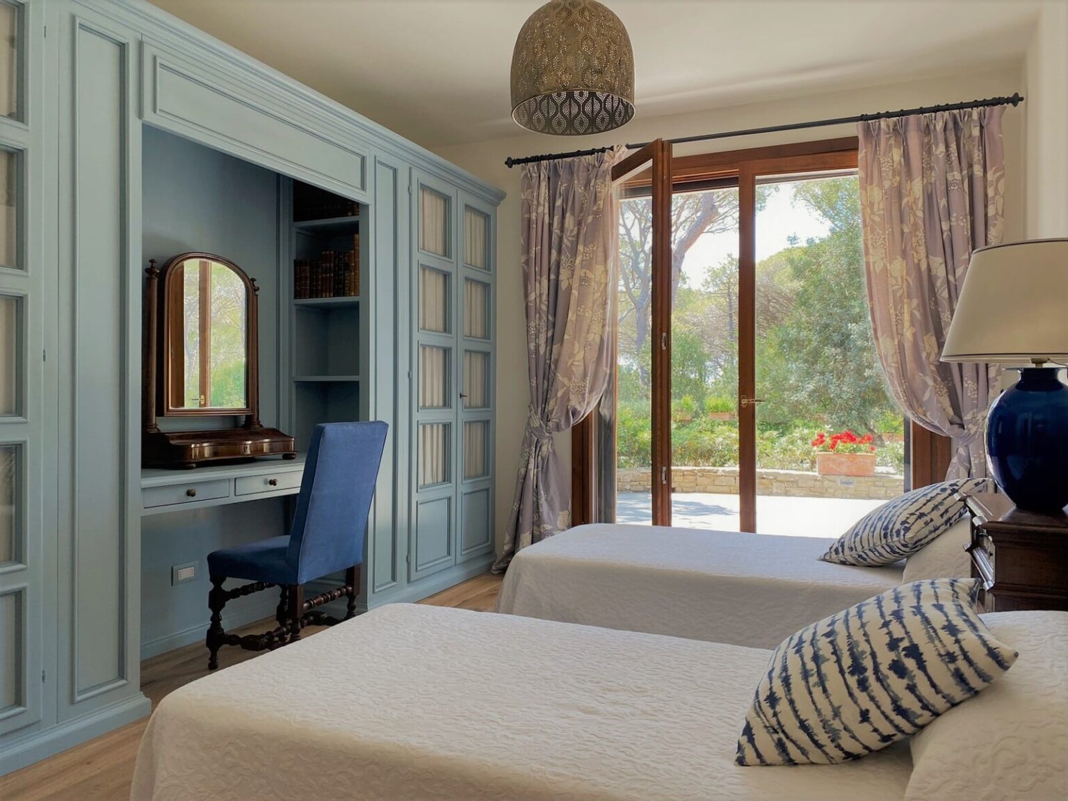 Cozy bedroom featuring two twin beds, a blue accent chair, a stylish vanity with a mirror, and large windows with sheer curtains overlooking a garden. The room has a light blue color scheme and elegant decor, creating a serene atmosphere. | Only on SopranoVillas