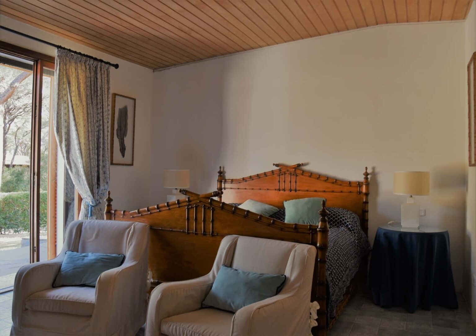 Cozy bedroom interior featuring a wooden bed with decorative headboard, two armchairs with cushions, and a bedside table. Natural light streams in through a large window with curtains, creating a warm and inviting atmosphere. | Only on SopranoVillas