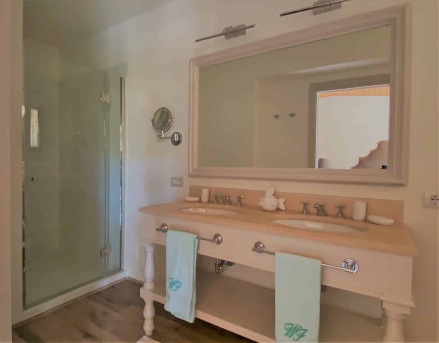 Modern bathroom featuring a double sink vanity with elegant fixtures, a large mirror, and a glass shower enclosure. Soft color palette with light wood accents and neatly placed towels. | Only on SopranoVillas