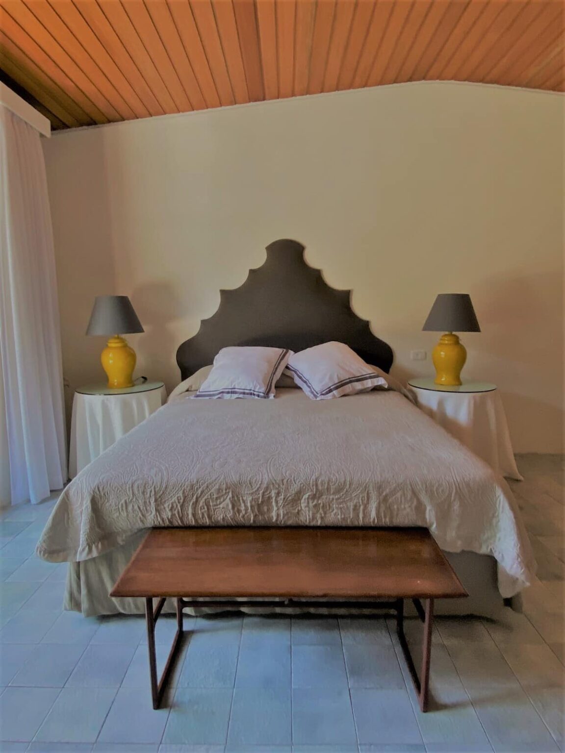Cozy bedroom featuring a king-sized bed with decorative pillows, two bedside lamps, and a wooden bench. The room has a warm color palette with wooden ceiling beams and soft lighting, creating a relaxing atmosphere. Ideal for vacation rentals or home décor inspiration. | Only on SopranoVillas