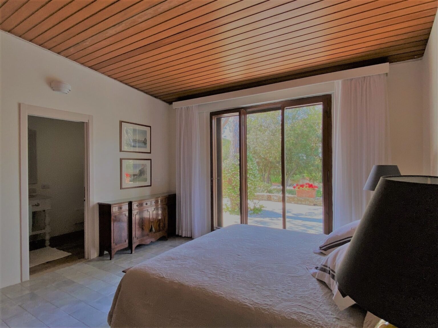 Cozy bedroom featuring a wooden ceiling, large sliding glass doors with views of a garden, and elegant decor including a bedside lamp and framed artwork. The room has a serene ambiance, perfect for relaxation and comfort. | Only on SopranoVillas