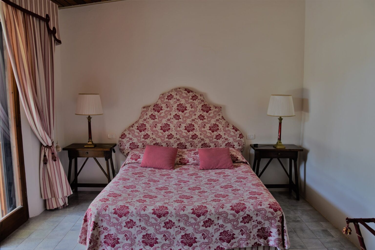 Cozy bedroom featuring a floral patterned bedspread, two pink cushions, bedside lamps, and large windows with sheer curtains, creating a warm and inviting atmosphere. | Only on SopranoVillas