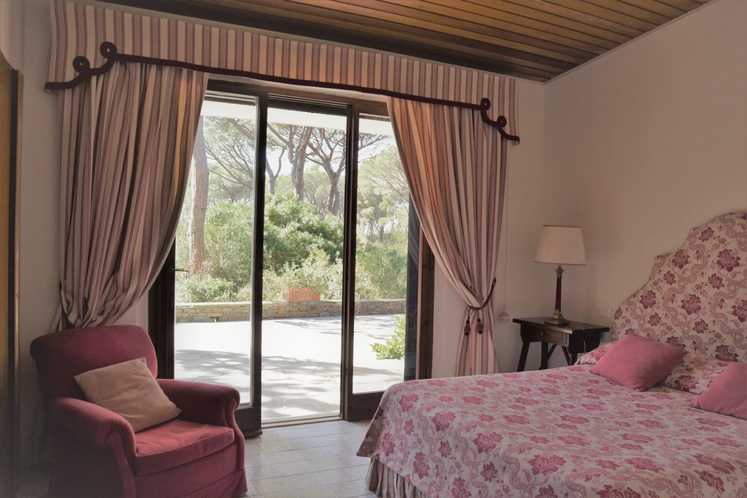 Cozy bedroom with floral bedding and draped curtains, featuring a view of a serene outdoor garden through sliding glass doors. A pink armchair and a bedside lamp add to the inviting atmosphere. | Only on SopranoVillas