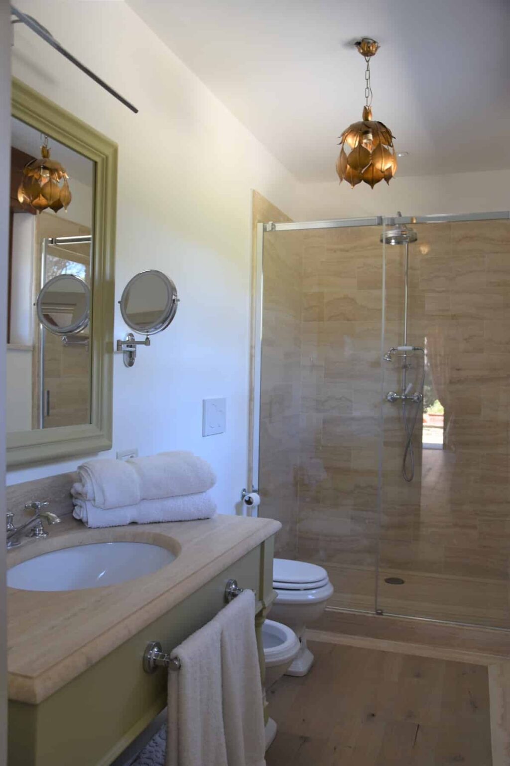 Modern bathroom interior featuring a glass shower, a wooden vanity with a sink, neatly stacked white towels, and decorative lighting. The design includes a green-framed mirror and a clean, minimalist aesthetic. | Only on SopranoVillas