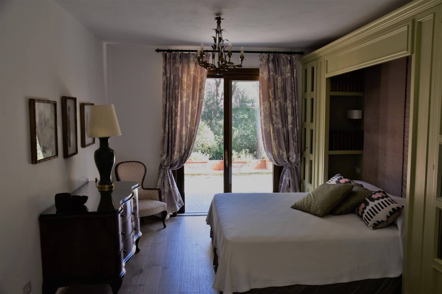 Cozy bedroom featuring a queen-sized bed with decorative pillows, a stylish chandelier, an elegant armchair, and large windows with curtains, offering a view of the outdoors. | Only on SopranoVillas