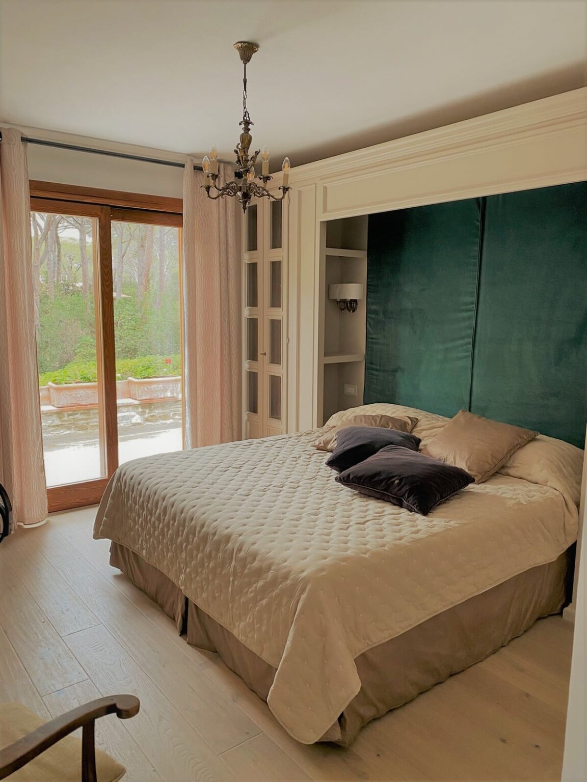 Cozy bedroom featuring a queen-sized bed with a beige quilt and decorative pillows, elegant chandelier lighting, and large windows with soft curtains overlooking a green outdoor space. The room has built-in shelves and a calming teal accent wall, creating a serene atmosphere. | Only on SopranoVillas