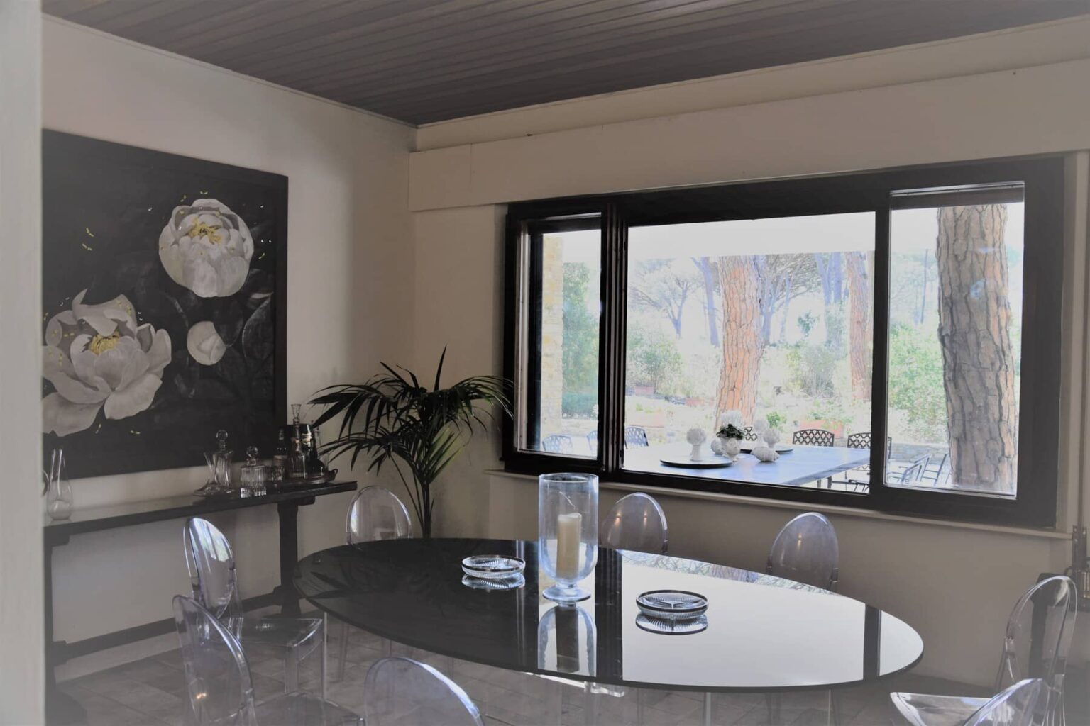 Modern dining room featuring a dark circular table, transparent chairs, and a large window showcasing a scenic outdoor view. A contemporary art piece with floral designs adorns the wall, complemented by indoor plants. | Only on SopranoVillas