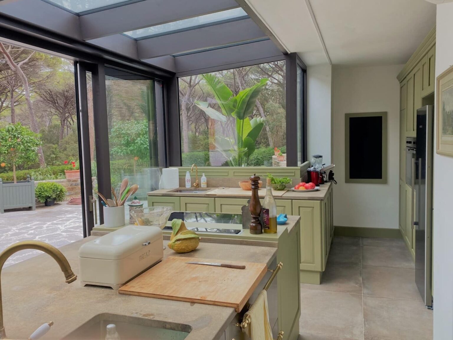 Modern kitchen with large windows overlooking a garden, featuring light green cabinetry, a wooden countertop, and kitchen appliances. The space is bright and airy, emphasizing a connection to nature. | Only on SopranoVillas