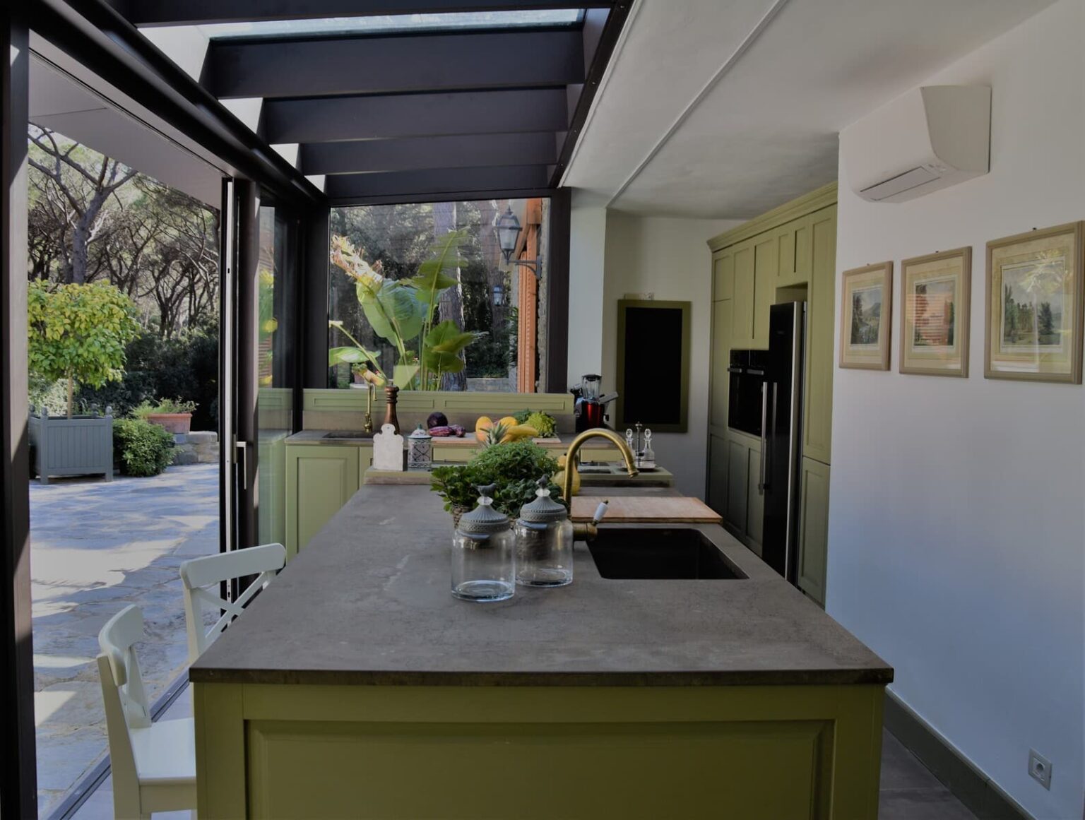 Modern kitchen with green cabinetry, a large island featuring a stone countertop, and a view of the outdoors through glass walls. The kitchen is well-lit and includes decorative plants, kitchen utensils, and framed artwork on the walls. | Only on SopranoVillas