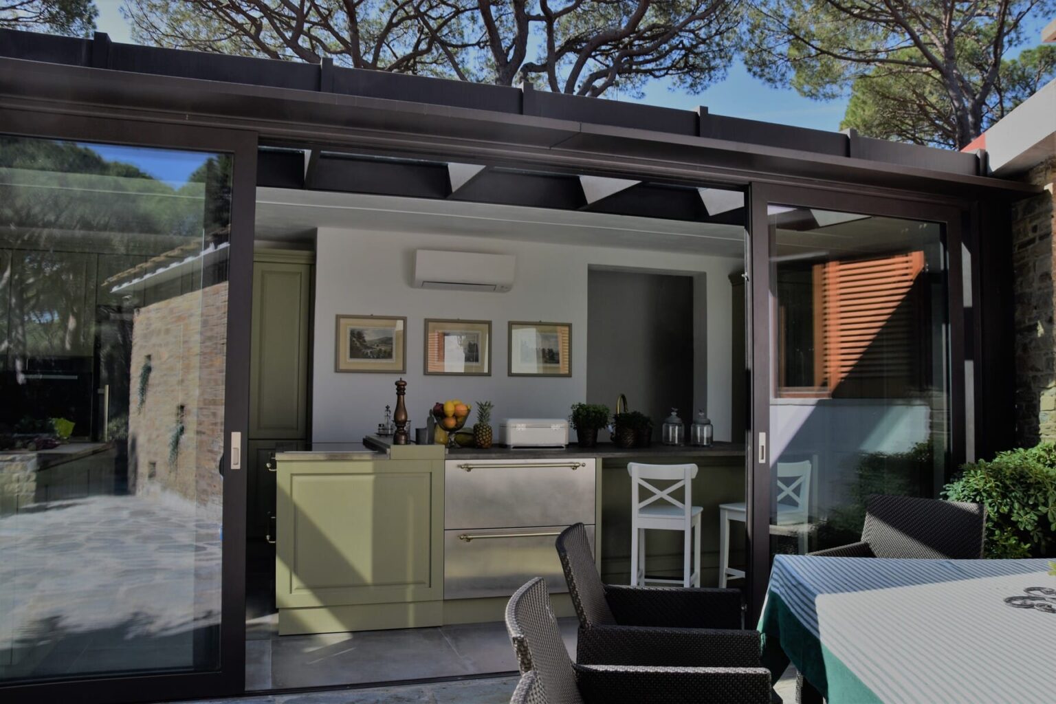 Modern kitchen with green cabinetry and stainless steel appliances, featuring a glass wall that opens to an outdoor dining area, surrounded by lush greenery and natural light. | Only on SopranoVillas