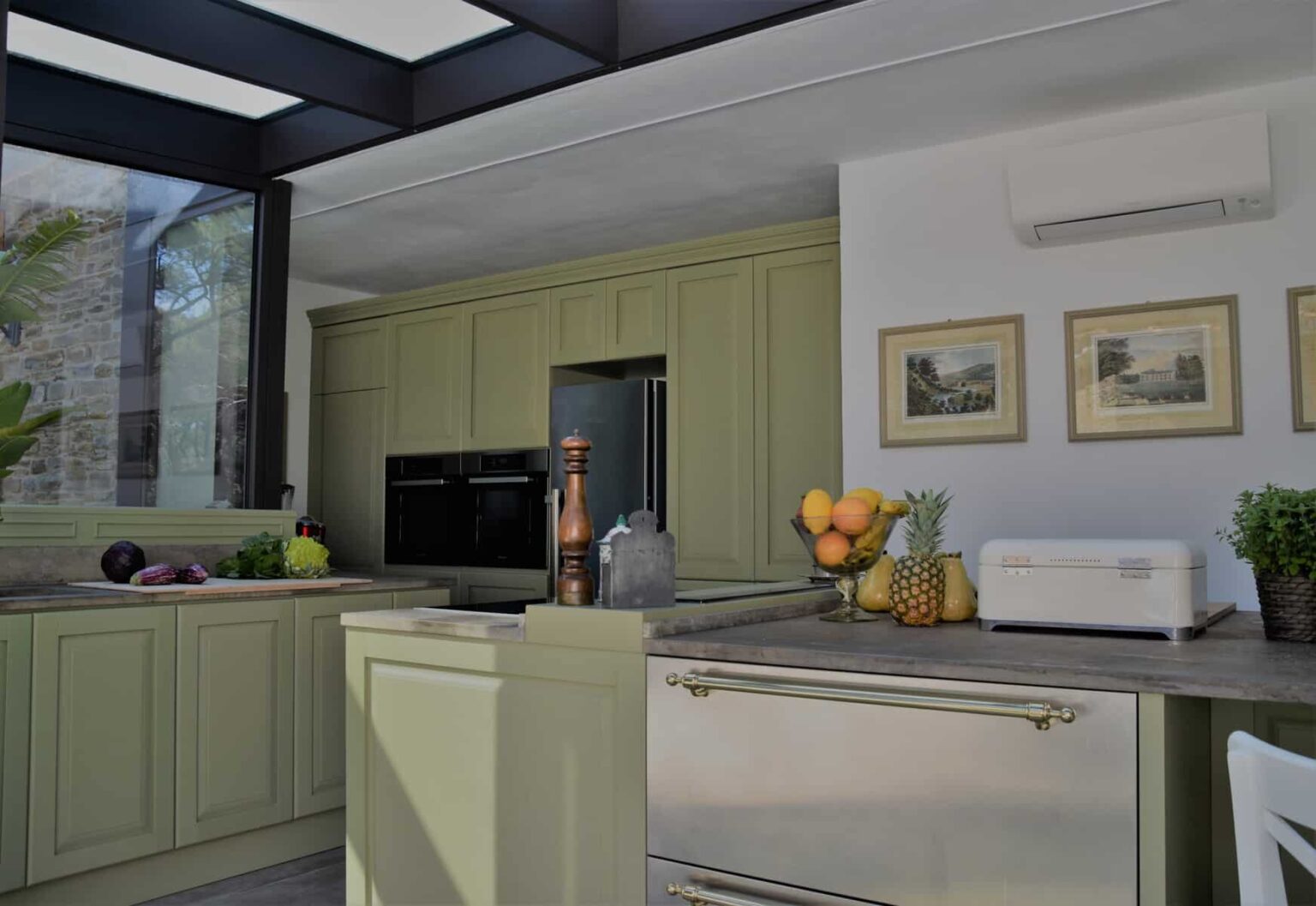 Modern kitchen with green cabinetry, stylish countertop, and stainless steel appliances, featuring fresh fruits and decorative artwork, illuminated by natural light from large windows. | Only on SopranoVillas