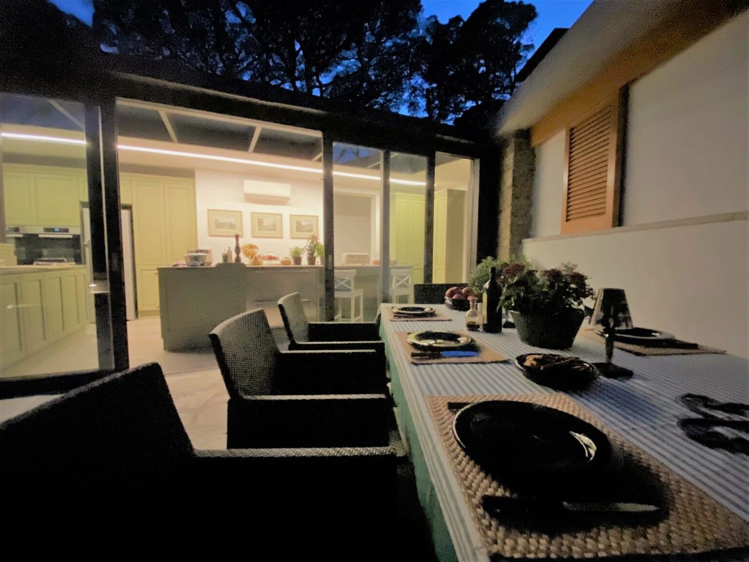 Cozy outdoor dining setup featuring a table with plates and a centerpiece, surrounded by dark wicker chairs, with a modern kitchen visible through large glass doors in the background, illuminated by soft evening light. | Only on SopranoVillas