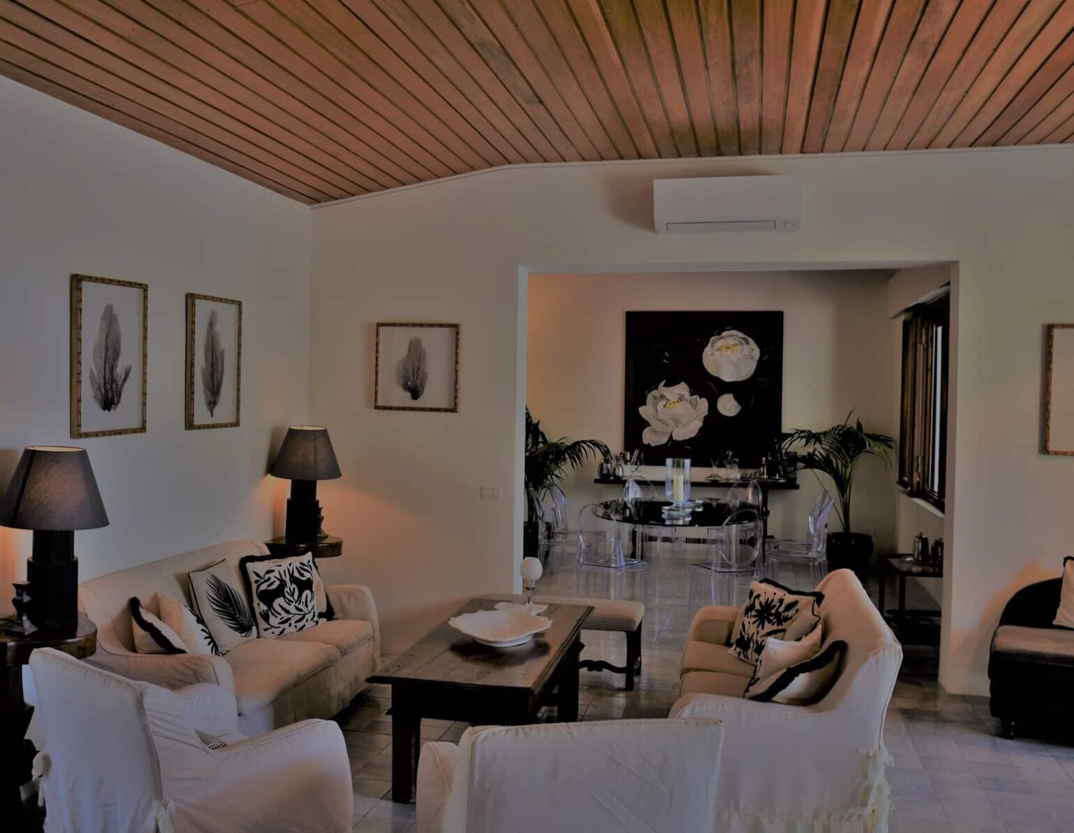 Modern living room interior featuring a cozy seating area with light-colored sofas, decorative pillows, and stylish lamps. The room includes framed botanical artwork, a wooden coffee table, and a dining area with a glass table. Elegant design elements and natural light create a welcoming atmosphere. | Only on SopranoVillas