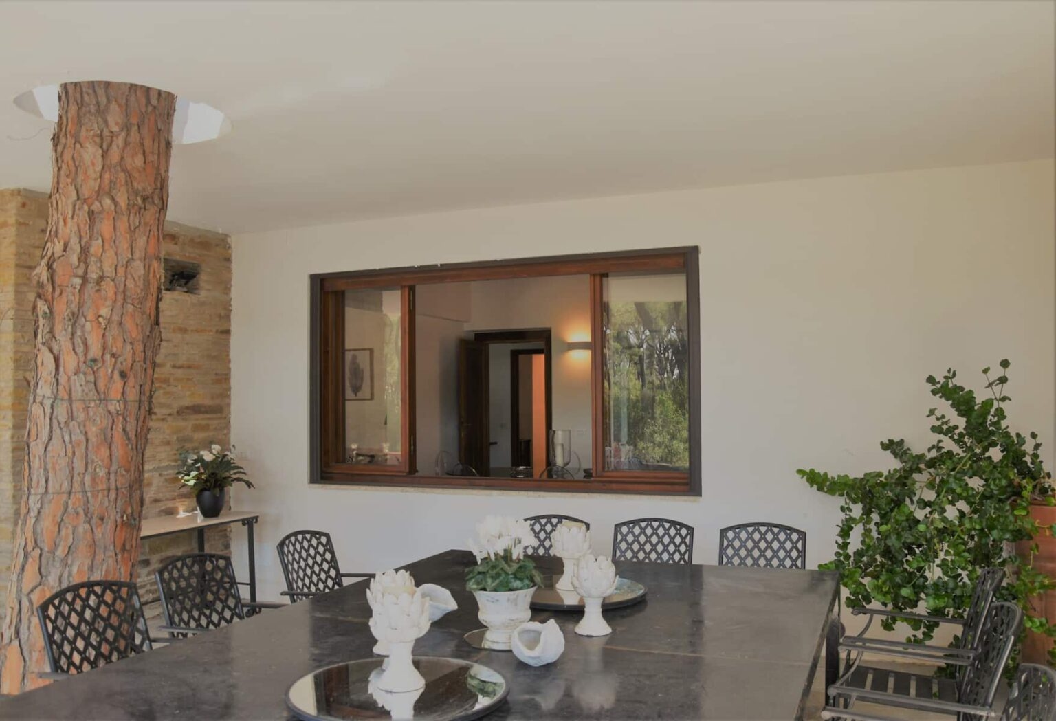 Modern dining area featuring a large table surrounded by black metal chairs, with a view of an outdoor space through a large window. Decorated with potted plants and a centerpiece, the space combines indoor and outdoor elements for a stylish dining experience. | Only on SopranoVillas