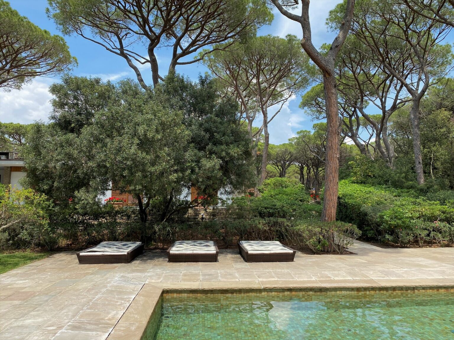 Lush outdoor space featuring three lounge chairs beside a serene swimming pool, surrounded by greenery and tall trees under a clear blue sky. | Only on SopranoVillas