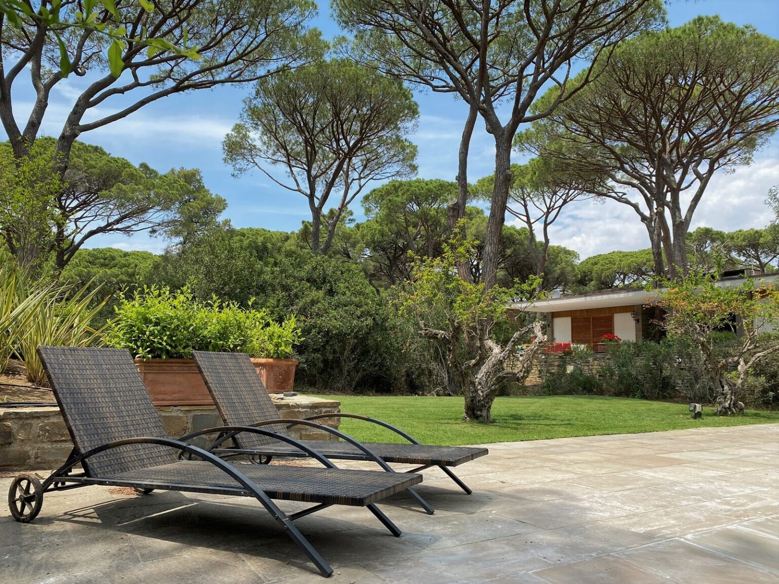 Two stylish lounge chairs positioned on a patio surrounded by lush greenery and tall trees, with a serene home in the background under a clear blue sky. | Only on SopranoVillas