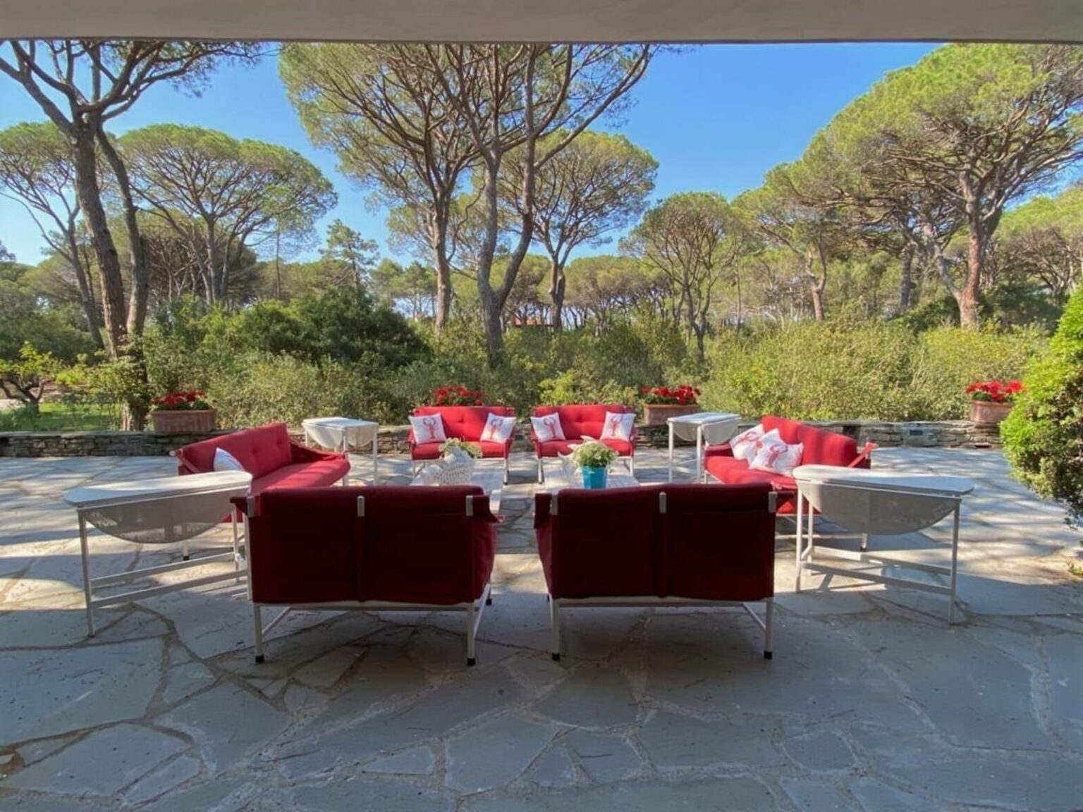 Outdoor seating area featuring red sofas and white tables, surrounded by lush greenery and tall pine trees, creating a serene and inviting atmosphere for relaxation and gatherings. | Only on SopranoVillas