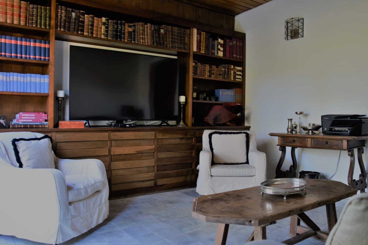 Cozy living room featuring a wooden bookshelf filled with books, a large flat-screen TV, two white armchairs, and a wooden coffee table. The space is well-lit and combines modern and classic decor elements. | Only on SopranoVillas