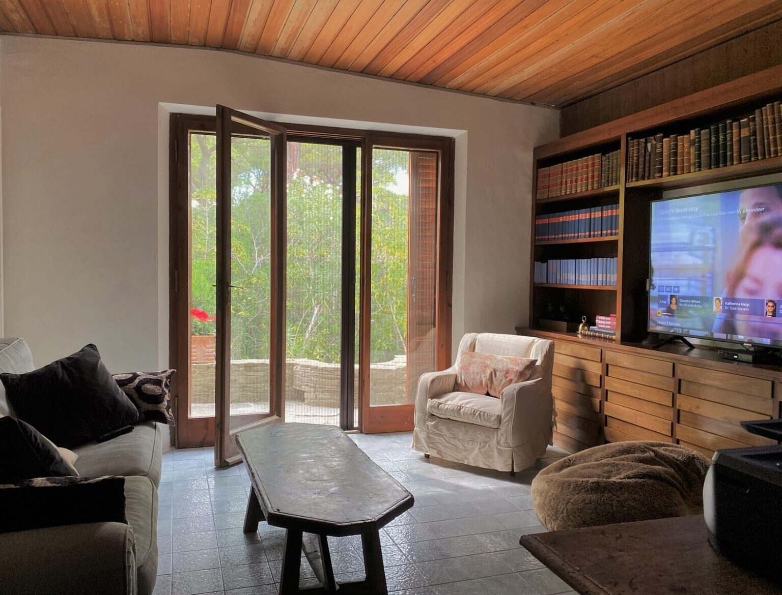 Cozy living room featuring a comfortable sofa with decorative pillows, a wooden coffee table, and a plush armchair. Large windows open to a lush outdoor view, while a TV displays content beside a bookshelf filled with books. The warm wooden ceiling adds a touch of elegance to the space. | Only on SopranoVillas
