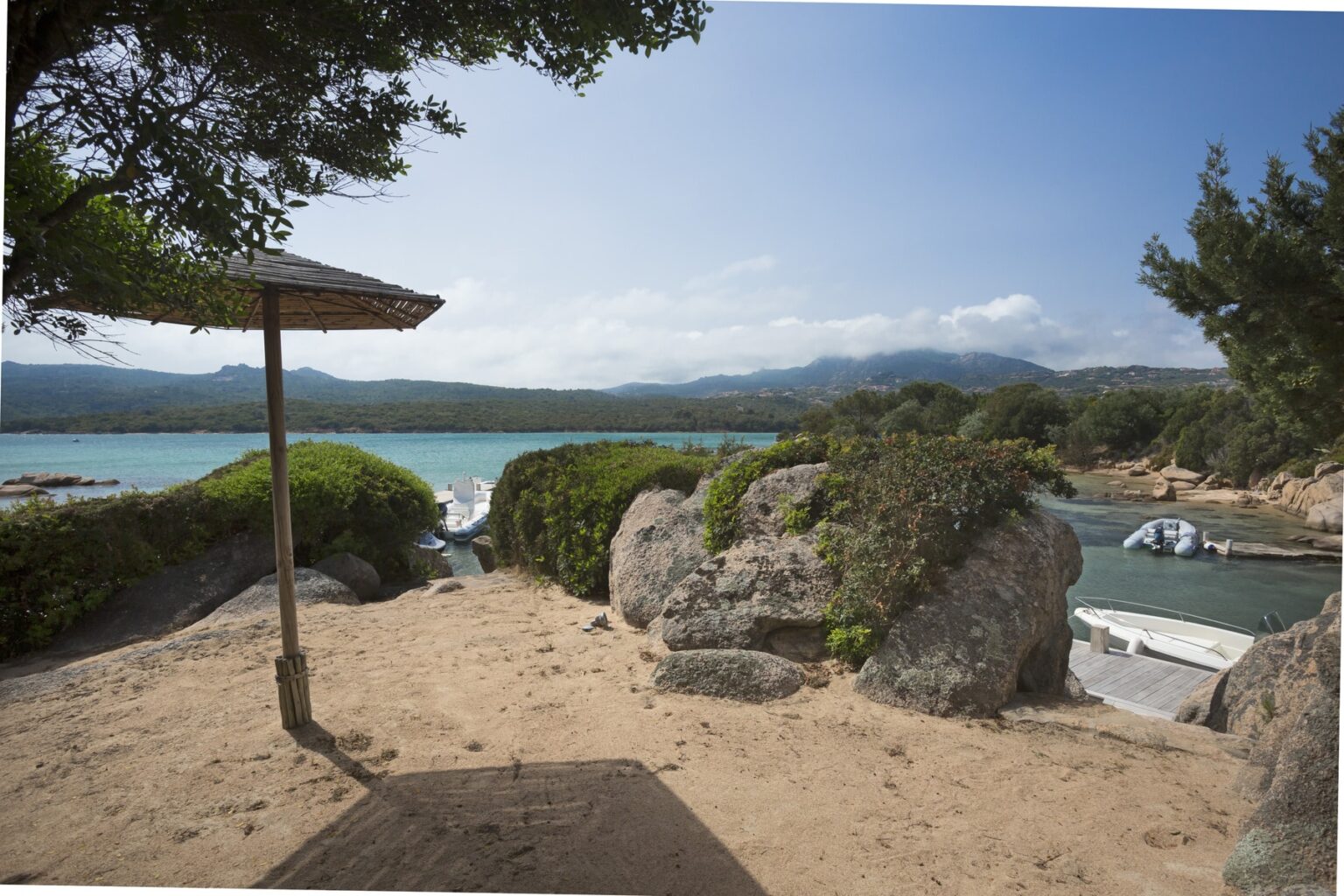 Scenic view of a sandy beach with a thatched umbrella, overlooking a tranquil turquoise bay surrounded by lush greenery and rocky formations, with boats anchored in the distance and mountains in the background. | Only on SopranoVillas