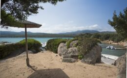 Scenic view of a sandy beach with a thatched umbrella, overlooking a tranquil turquoise bay surrounded by lush greenery and rocky formations, with boats anchored in the distance and mountains in the background.